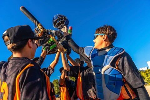 Young baseball players huddling together, lifting catcher's gear and a bat into the bright blue