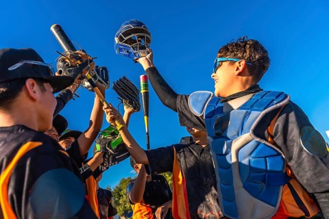 Young baseball team members cheering together, raising bats, gloves, and a catcher's helmet into