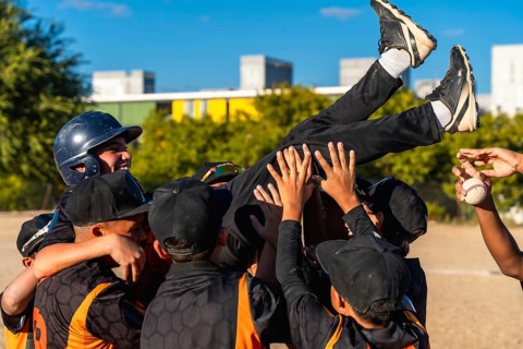 Young baseball teammates lift a smiling boy in celebration after a game, sharing joyful camaraderie