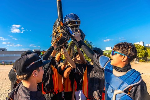 Baseball players huddling together, raising their catcher's helmet, bat, and gloves in the air,