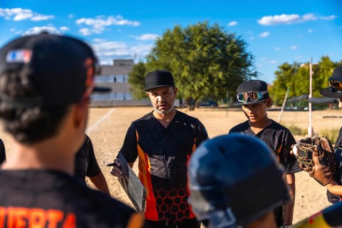 Baseball coach in a black and orange jersey and cap huddling with his youth team, discussing game