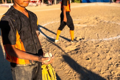 Young baseball boy in orange and black uniform holds glove and ball on sunlit dirt field during