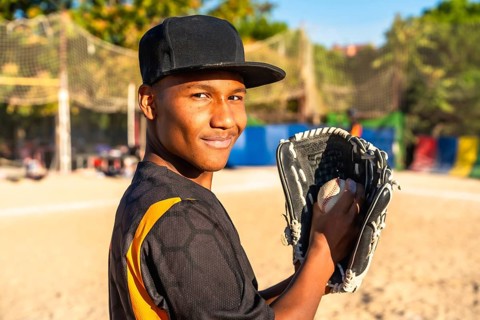 Young male baseball player stands on a sunny field, smiling confidently while holding a baseball in