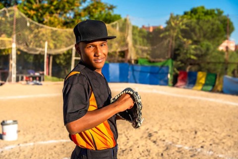 Confident young baseball player stands on dusty field gripping his glove, smiling at the camera and