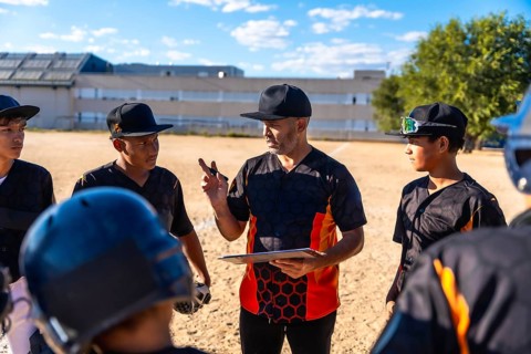 Baseball coach in a uniform and cap holding a clipboard, explaining strategy to a group of diverse