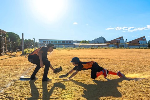 Kids playing baseball on a dusty field, one player sliding into home plate kicking up dust while