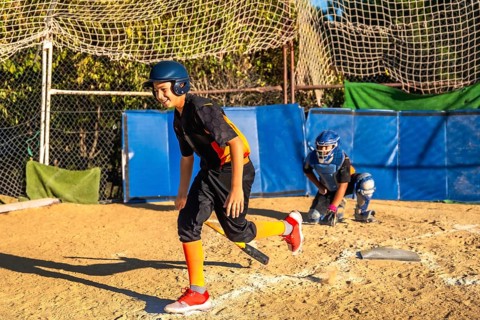 Young boy smiles while running on the dirt field, holding a bat and wearing a helmet and uniform,