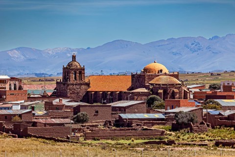 Historic church in a village with brick red roofs against a mountain backdrop under clear skies,