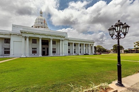 A white building with dome and pillars surrounded by green lawn under cloudy sky, Colombo Sri Lanka