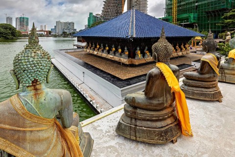 Buddha statues with yellow cloths sit in front of a modern cityscape on the water, The Seema Malaka