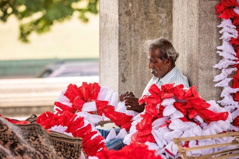 Elderly man arranges red and white flowers under a stone pillar in a warm, calm atmosphere, people