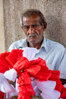 Close-up of an elderly man with red and white decorative flowers in the foreground, people making