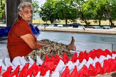 Elderly woman sitting outdoors working on red and white decorations, people making garlands in
