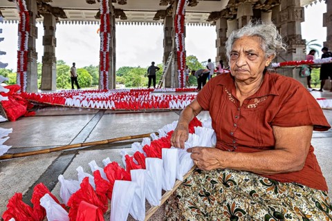 Elderly woman sitting and making red and white decorations in a hall, people making garlands in
