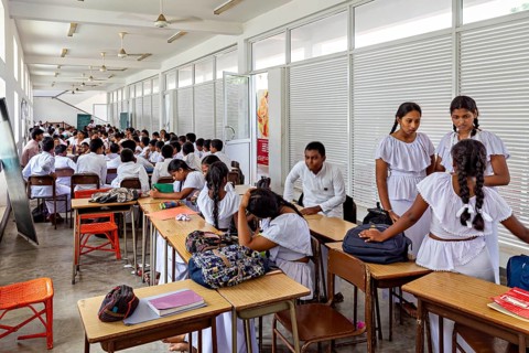 Students in uniforms sit in a bright classroom, some talk while others study, young people in a