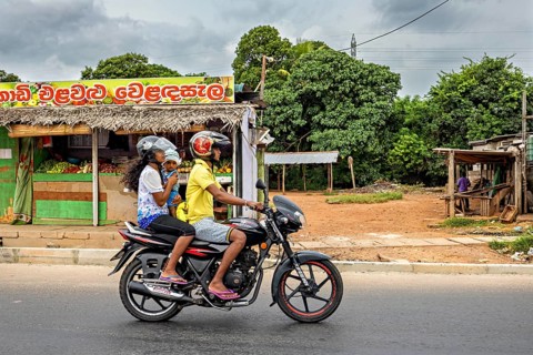 Two people on a motor scooter ride down a street, past a small shop and tropical plants, a family