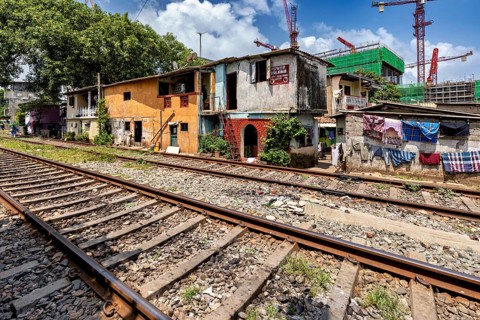 Construction cranes rise behind brightly painted houses along the train tracks under a blue sky,
