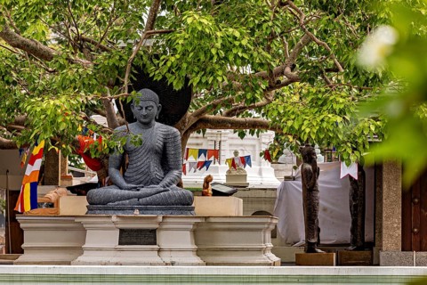 Stone Buddha under trees with decorative prayer flags in the temple area, The Seema Malaka Temple