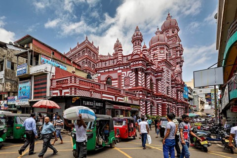 Bustling street scene with rickshaws and passers-by in front of a red architectural building, the