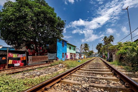 Railway tracks run along colorful houses and a large tree under a cloudy sky, railway line in the