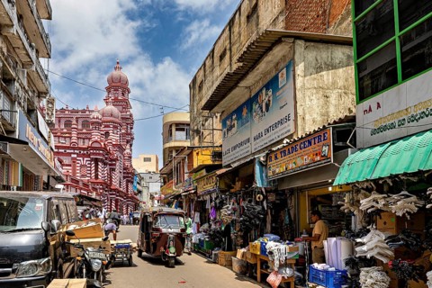 Bustling market street with people and stalls, red architectural building in the background, the