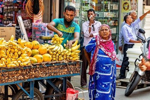 A busy street market with a fruit vendor selling bananas and papayas. A woman in colorful clothes