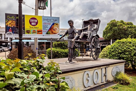 Sculpture of a person drawing a rickshaw surrounded by green bushes and advertising in the