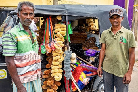 Two men stand in front of a tuk-tuk sales stand stocked with sponges on a busy street, tuk tuk as a
