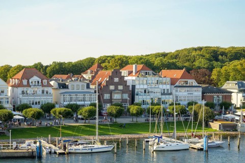 Picturesque houses and boats stand on a sunny harbor against a green forest backdrop,