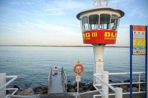 A modern DLRG watchtower looks out at the vast water while a bather sits on the jetty,