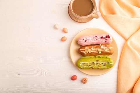 Glazed Eclairs on white wooden background and orange linen textile, cup of coffee, top view, flat