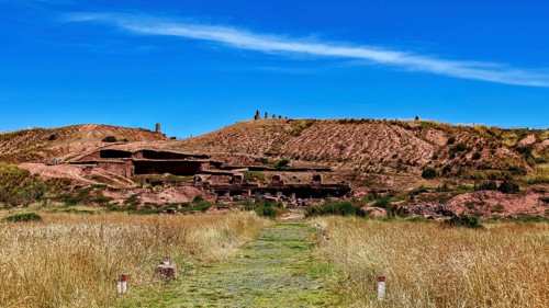 Grassy trail leads to ancient ruins framed by natural landscape under blue skies, the