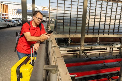 Solo male traveler waiting at a modern railway station, holding a yellow suitcase and checking his
