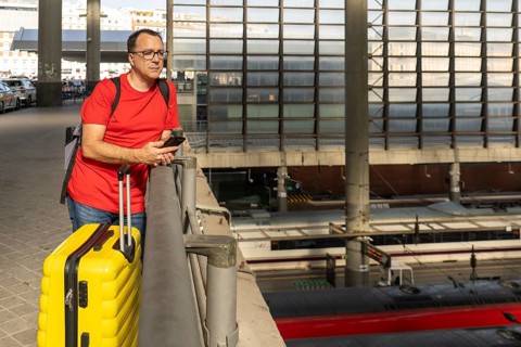 Adult male traveler waiting at an elevated railway station, holding a yellow suitcase while