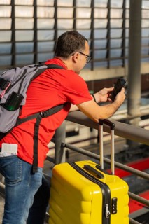 Mature male traveler using a smartphone at a train station while standing next to a yellow suitcase