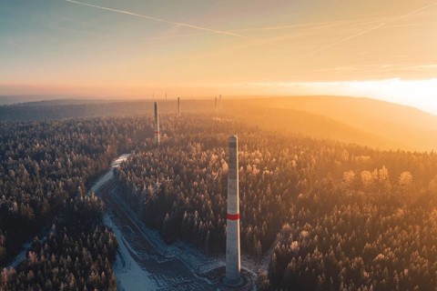 Sun rays over a foggy forest and wind turbine towers rising from the tree cover, Windkraft Bau,