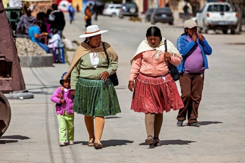 Two woman and a little girl walk down a busy street in traditional clothes, people in the altiplano