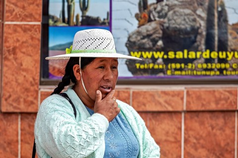 Woman in traditional dress wearing hat on the street in front of a tiled wall with advertising,