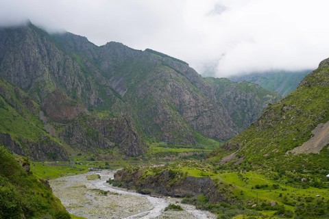 Mighty mountains with cloud cover, a river cuts through the green valley with rocks and vegetation,