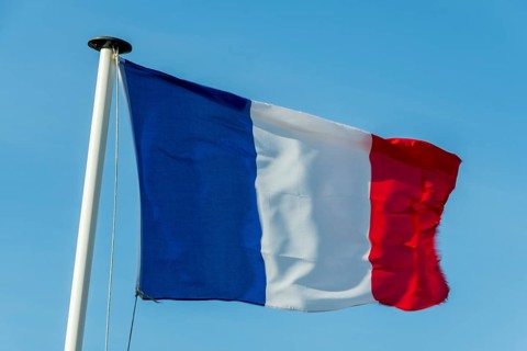 A French flag flutters in the wind on a white flagpole. The background is a clear, azure sky