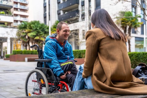 Young man in a wheelchair smiling and talking with a woman outdoors in an urban setting,