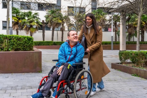 Caregiver pushing a man in a wheelchair on a paved path in an urban park, sharing a happy moment