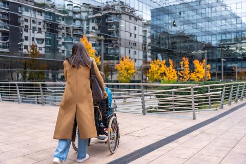 Woman in a brown coat pushing a young person in a wheelchair along a modern urban sidewalk,