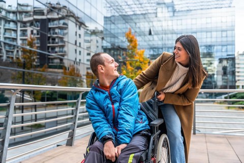 Caregiver woman pushes smiling young man in wheelchair along a city sidewalk, sharing a warm moment