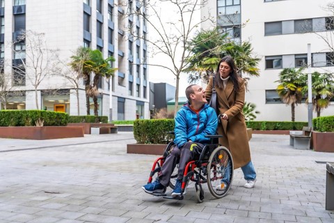 Woman pushing smiling man in a wheelchair along a city sidewalk, conveying everyday support,