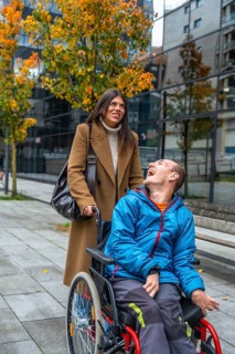 Woman pushes cheerful man in a wheelchair along a city sidewalk lined with autumn trees, sharing