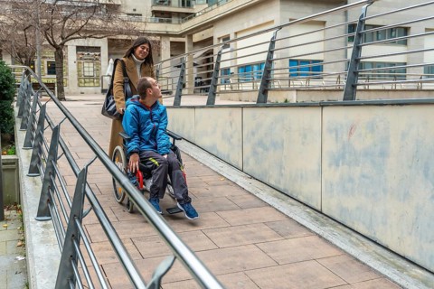 Woman pushing a young man with a disability in a wheelchair, navigating an accessible ramp with