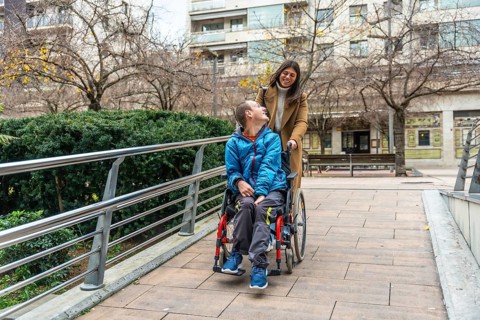 Caretaker pushing young man in wheelchair along accessible urban path, both smiling and sharing a