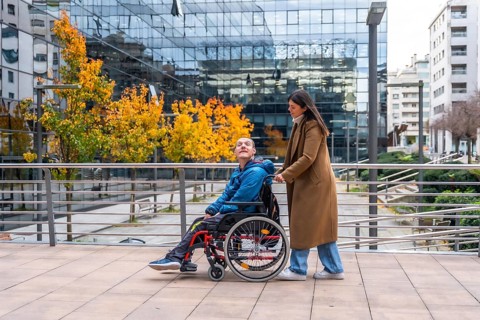 Woman pushing a smiling man in a wheelchair along an urban sidewalk during autumn, showcasing