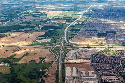 An aerial view shows a vast landscape of highways, fields and densely built suburbs under clouds,
